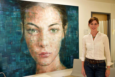 Beschäftigte in weißer Bluse steht vor einem riesen-großen Gemälde, auf dem das Gesicht einer Frau zu sehen ist.
