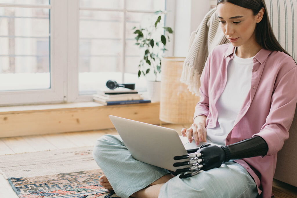 Frau mit Armprothese sitzt mit Laptop auf dem Boden und lächelt in Bildschirm