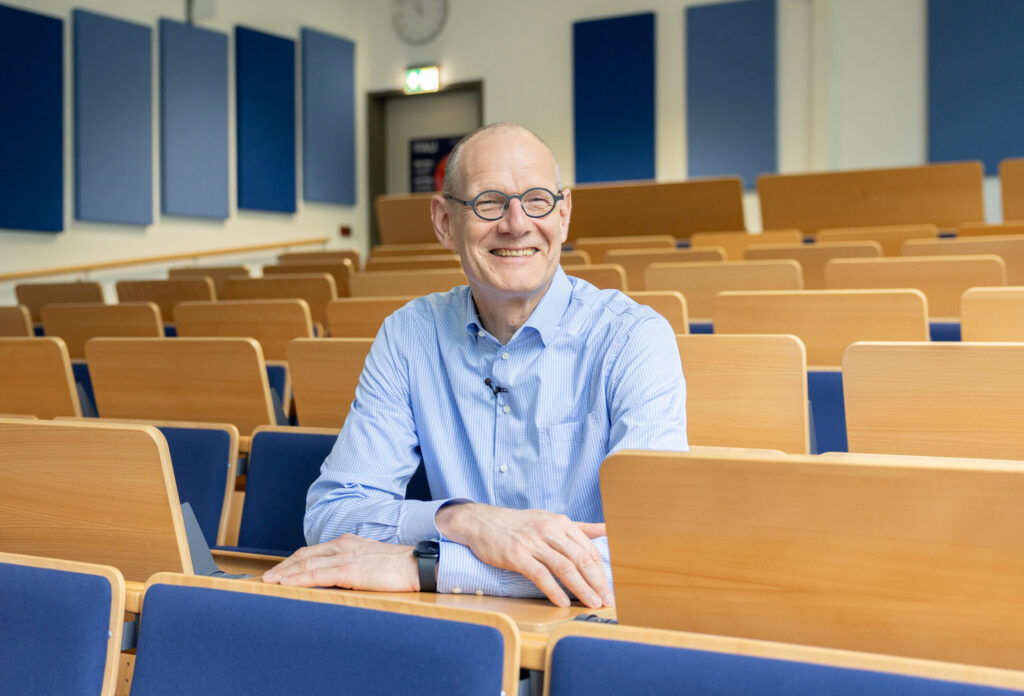 Bernd Montag sitzt lächelnd in einem Hörsaal des Physikums der FAU auf einem der Holzklappstühle mit blauem Sitzbezug. Es sind keine weiteren Personen sichtbar.