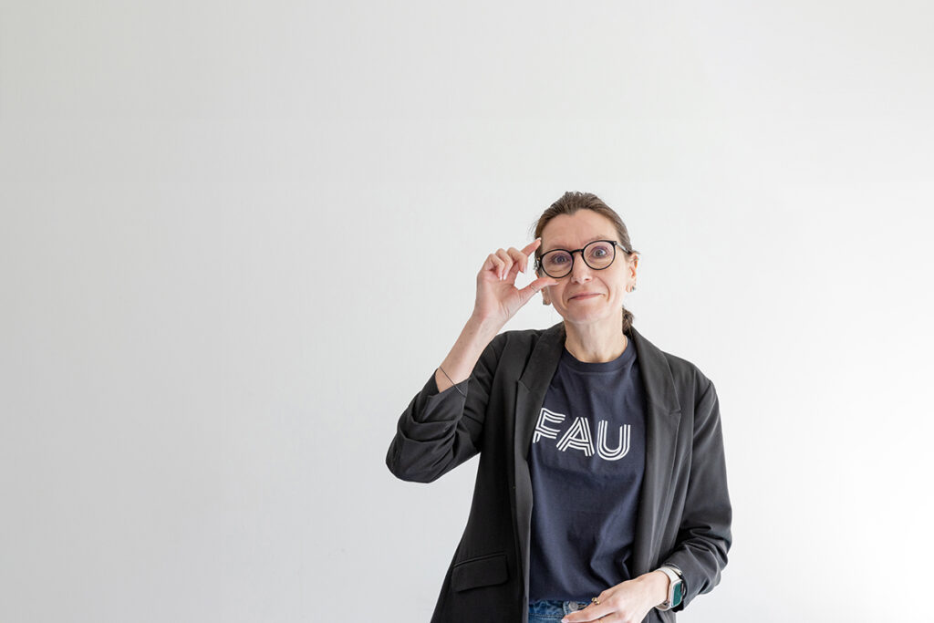 Prof. Dr. Aline Bozec steht vor einer weißen Wand und fasst sich an die Brille. Sie trägt ein T-Shirt mit FAU-Logo.