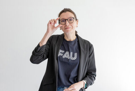 Prof. Dr. Aline Bozec steht vor einer weißen Wand und fasst sich an die Brille. Sie trägt ein T-Shirt mit FAU-Logo.