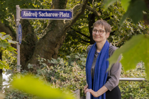 Prof. Dr. Eva Pils blickt an der Kamera vorbei und lächelt dezent. Hinter ihr sind Bäume und ein Straßenschild des "Andreij-Sacharow-Platz".