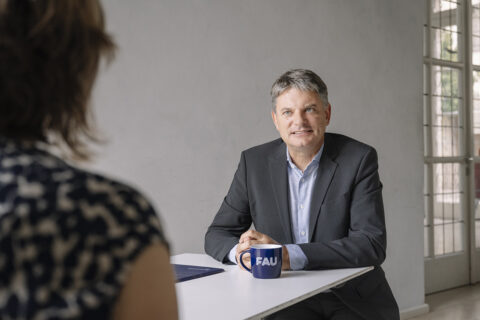 Das Bild zeigt den FAU-Präsidenten an einem Tisch sitzend. Er unterhält sich mit einer ihm gegenüber sitzenden Frau. Vor ihm ist eine Tasse mit FAU-Logo platziert.