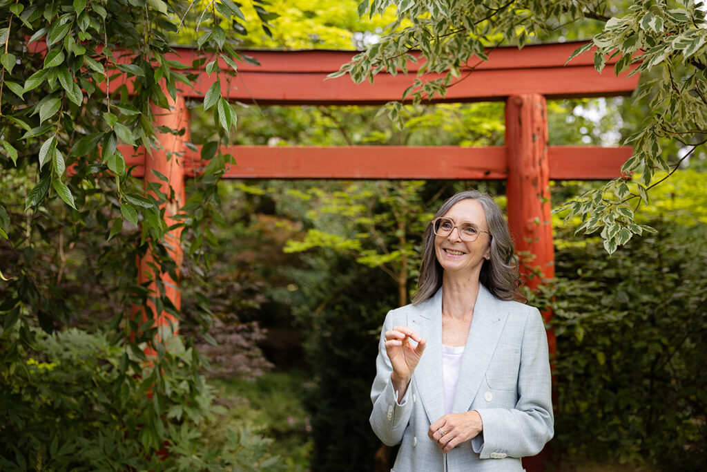 Andrea Bréard befindet sich in einem Garten mit einem orange-braunem, hölzernen Tor im Hintergrund.