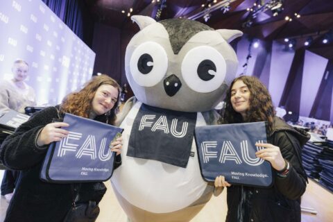 Zwei Studentinnen und die FAU-Eule. Die Studentinnen halten ihre FAU-Taschen hoch.