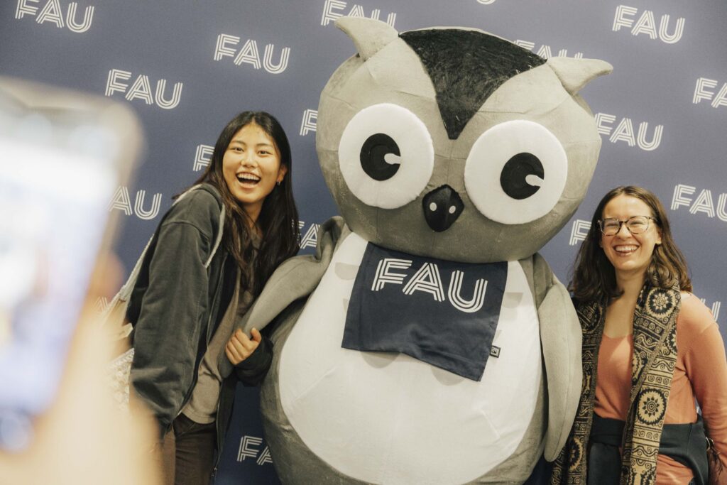 Zwei Studentinnen mit FAU-Eule. Sie werden mit einem Handy fotografiert.