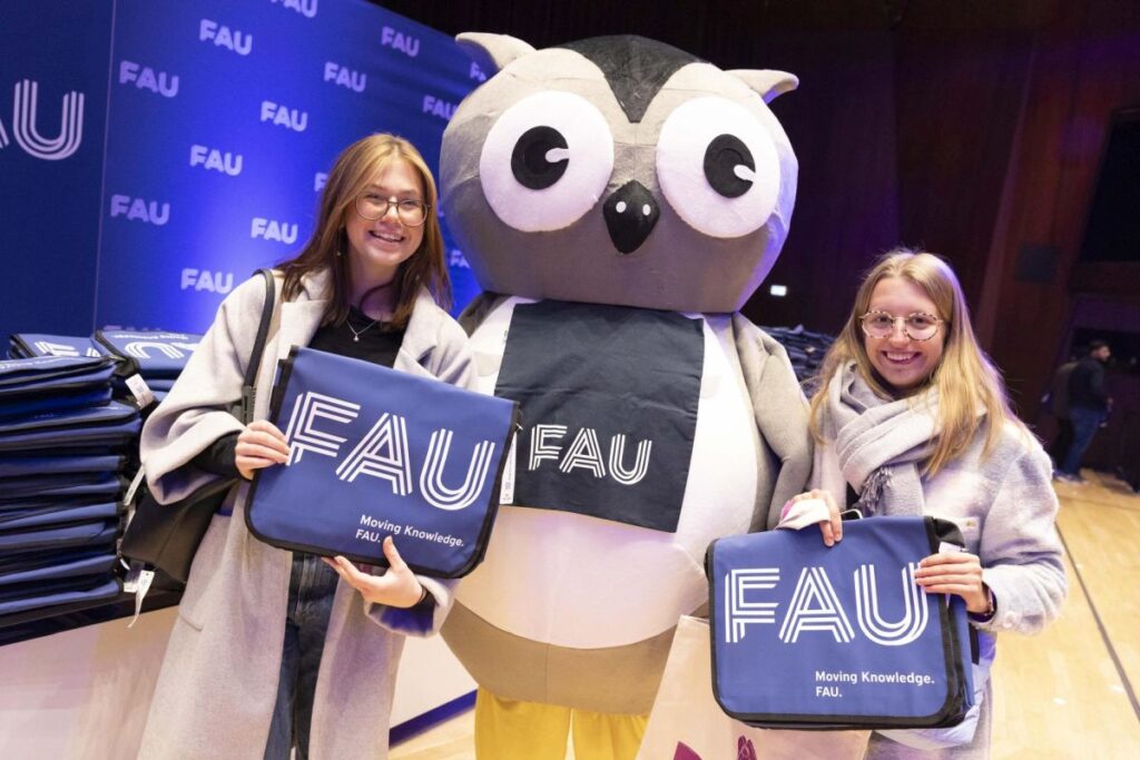 Erstsemesterbegrüßung 2023 der Friedrich-Alexander-Universität Erlangen-Nürnberg (FAU), 2 Studentinnen mit FAU-gebrandetetn Taschen lächeln neben einer Person in einem Eulen-Kostüm in die Kamera