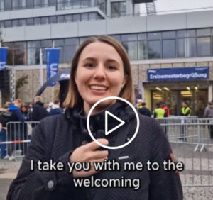 A woman holds a microphone in front of the Heinrich-Lades-Halle in Erlangen where the new students’ welcome event took place on October 14. People in the background and FAU flags.