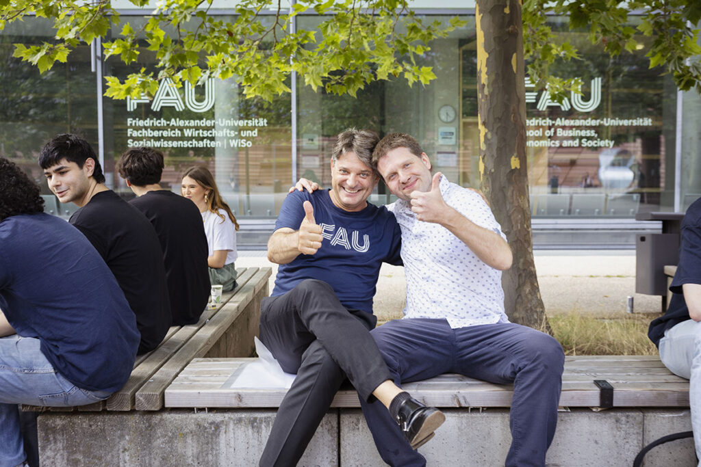 Hornegger and Hechtner are sitting together on a bench in front of a building at the School of Business, Economics and Society and showing a thumbs up.