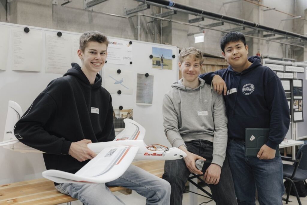 Drei Jugendliche; einer hält ein Modellflugzeug in der Hand, im HIntergrund ein Whiteboard an dem Skizzen und Fotos befestigt sind.