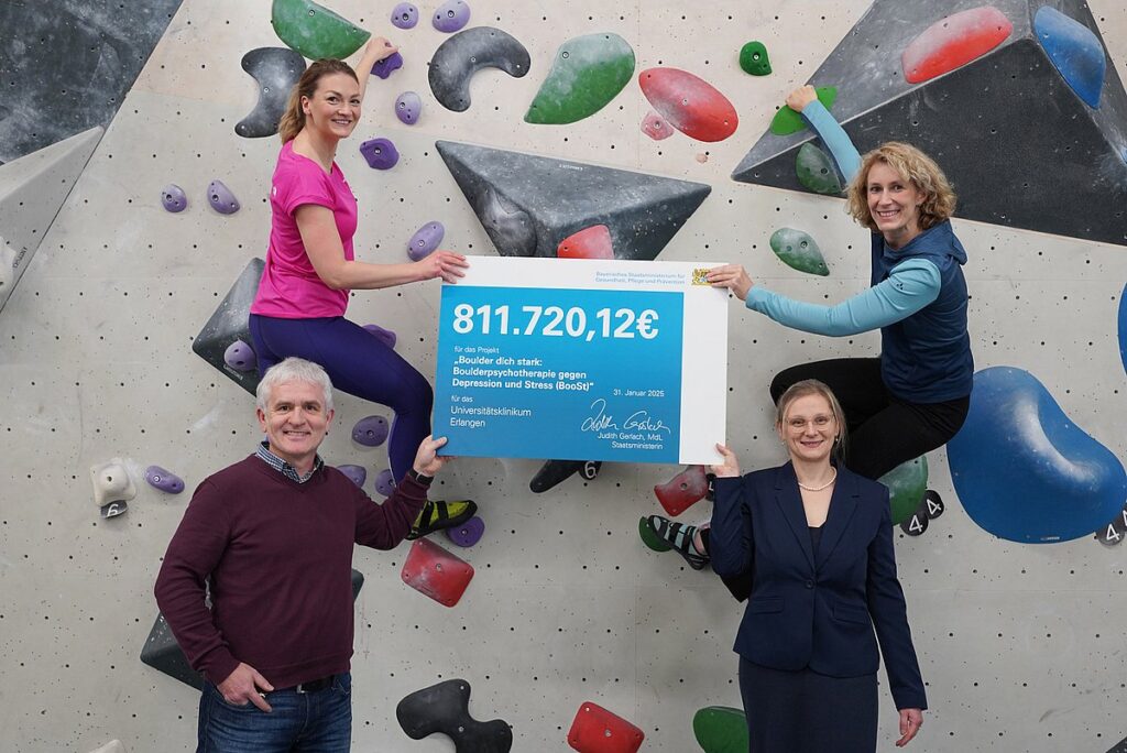 Akteure mit einem Scheck an der Hand. Ministerin Gerlach und Katharina Luttenberger hängen an einer Boulderwand, darunter stehen Studienleiterin Prof. Dr. Carolin Donath (u. r.) und Prof. Dr. Oliver Kratz.